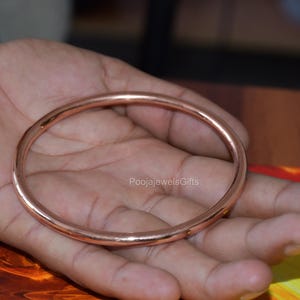 Könnte beinhalten: Ein glatter, kreisförmiger Kupferarmreif mit einer polierten, reflektierenden metallischen Oberfläche. Das Armband zeichnet sich durch ein schlichtes, durchgehendes Design mit gleichmäßiger Dicke aus und zeigt einen warmen, rotbraunen Metallton.