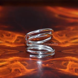 Könnte beinhalten: Ein silberfarbener Ring mit einem durchgehenden Spiraldesign, das sich mehrfach um sich selbst wickelt. Die Metalloberfläche ist glatt und reflektierend und weist ein poliertes Finish auf. Der Text "PoojajewelsGifts" ist auf dem Ringband zu sehen.