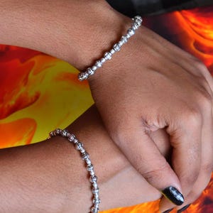 Könnte beinhalten: Zwei silberfarbene Armbänder mit einem sich wiederholenden Muster aus kleinen, kugelförmigen Perlen mit komplizierten, geätzten blumenartigen Details. Die Armbänder bestehen aus einer Reihe verbundener Metallsegmente, die ein strukturiertes, einheitliches Aussehen erzeugen.
