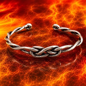 Könnte beinhalten: Ein silberfarbenes Metall-Armband mit einem gedrehten Drahtdesign, das ein zentrales Knotenmuster bildet. Das Armband hat eine offene Konstruktion mit abgerundeten Enden und zeigt eine polierte Oberfläche mit dunklen, oxidierten Akzenten.