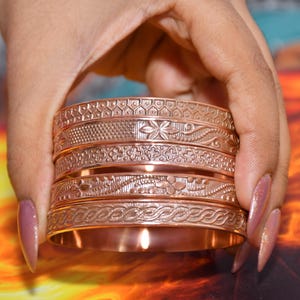May include: A set of four copper-toned metal bangles featuring intricate embossed patterns. Each bracelet displays a unique texture, including floral motifs, geometric shapes, and braided designs, all finished with a polished metallic sheen.
