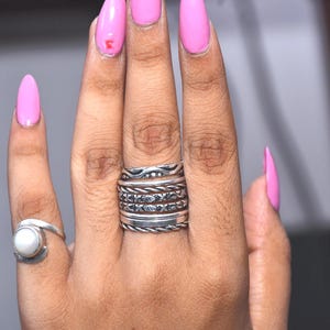 Könnte beinhalten: Ein Set aus silberfarbenen Ringen an zwei Fingern. Ein Finger zeigt einen Stapel aus fünf verschiedenen Ringen mit unterschiedlichen Texturen, darunter gedrehte Kordelmuster und Gravuren. Der andere Finger trägt einen Ring mit einem runden weißen Stein.