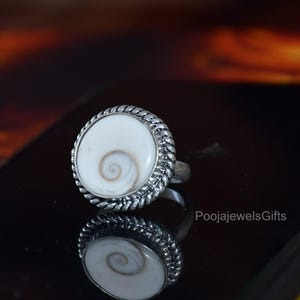 Könnte beinhalten: Ein Silberring mit einem runden, weißen Shiva-Auge-Muschelstein, der in der Mitte ein natürliches braunes Spiralmuster aufweist. Die Muschel ist in eine gedrehte silberne Seilfassung eingefasst und die Ringschiene ist schlicht. Der Text PoojaJewelsGifts ist sichtbar.