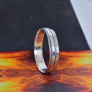 Könnte beinhalten: Ein silberfarbener Metallring mit einer glatten Innenschiene und einer strukturierten Außenschiene mit komplexen, geätzten Mustern. Der Ring hat eine polierte, reflektierende Oberfläche und ein abgerundetes Profil mit abwechslungsreicher Textur.
