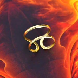 Könnte beinhalten: Ein goldfarbener Metallring mit offenem Bypass-Design und zwei überlappenden Bändern, die eine durchgehende, fließende Form bilden. Der Ring hat eine glatte, polierte Oberfläche und eine geschwungene, asymmetrische Silhouette.