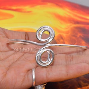 Könnte beinhalten: Ein silberfarbenes Metall-Armband mit zwei markanten, symmetrischen Spiralen in der Mitte. Das Band ist glatt und poliert, wobei sich die Spiralen in entgegengesetzte Richtungen drehen. Der Text "PoojajewelsGifts" ist auf dem Metall zu sehen.
