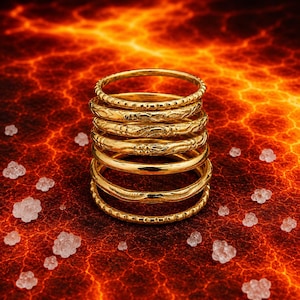 Könnte beinhalten: Ein Stapel aus sieben goldfarbenen Ringen mit unterschiedlichen Texturen und Mustern, darunter glatte, perlenartige und gravierte Designs. Die Ringe sind vertikal in einer einzigen Säule angeordnet und zeigen eine polierte metallische Oberfläche.