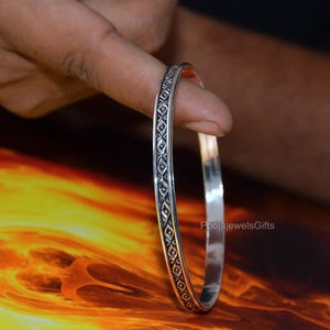 Könnte beinhalten: Ein silberner Armreif mit einem sich wiederholenden geometrischen Muster aus kleinen Rauten und Punkten, die in die Außenfläche eingraviert sind. Das Metall hat eine polierte Oberfläche mit abgedunkelten Vertiefungen, die das Design betonen.