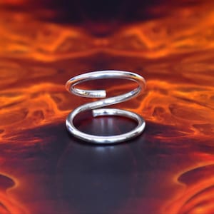 Könnte beinhalten: Ein silberfarbener Metallring mit einem Bypass-Design, das aus zwei offenen, sich überlappenden kreisförmigen Bändern besteht. Der Ring hat eine glatte, polierte Oberfläche und eine durchgehende, abgerundete Drahtstruktur.