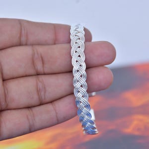 Könnte beinhalten: Ein silberfarbenes Armband in Manschettenform mit einem geflochtenen Drahtmuster. Das Armband besteht aus mehreren dünnen, miteinander verflochtenen Metallsträngen, die ein strukturiertes, seilartiges Erscheinungsbild über die gesamte Länge erzeugen.