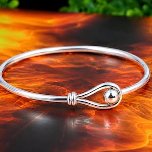 May include: A polished silver bangle bracelet featuring a smooth, circular band that terminates in a looped knot design. The knot is secured by a small, round silver bead nestled within the loop, creating a continuous, reflective metallic finish.
