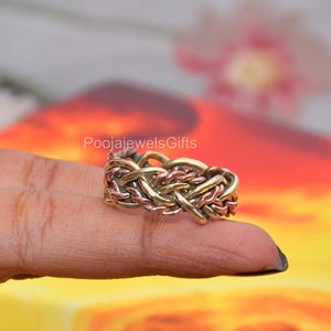 Könnte beinhalten: Ein mehrfarbiger Ring mit einem komplexen, geflochtenen Design aus abwechselnden Strängen aus poliertem goldfarbenem Metall und kupferfarbenem Metall. Das Band hat eine texturierte, geflochtene Optik mit dem Text PoojaJewelsGifts darüber.