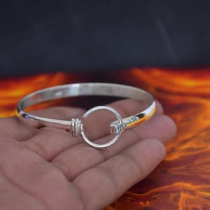 Könnte beinhalten: Ein polierter Silberarmreif mit einem kreisförmigen Ringverschlussmechanismus. Das Armband hat eine glatte, reflektierende metallische Oberfläche und ein durchgehendes Band, das mit einer zentralen kreisförmigen Schlaufe verbunden ist.