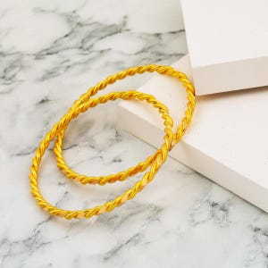 Könnte beinhalten: Zwei goldene Armreifen mit gedrehtem Seildesign. Die Armbänder sind aus poliertem gelbem Metall mit reflektierender Oberfläche gefertigt und weisen ein durchgehendes Spiralmuster auf, das für eine texturierte Optik am gesamten Band sorgt.