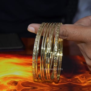 Könnte beinhalten: Ein Set aus fünf goldfarbenen Metallarmreifen. Die beiden äußeren Armreifen zeigen ein wiederkehrendes Blumenmuster, während die drei inneren Armreifen eine glatte, gehämmerte Textur aufweisen. Die Armreifen sind gestapelt und glänzend.