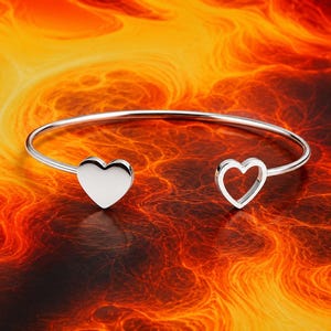 Könnte beinhalten: Ein silberfarbenes Armband in offener Form mit zwei herzförmigen Anhängern an den Enden. Ein Anhänger ist ein massives, poliertes Herz, während der andere ein offener Herzumriss ist. Das Armband hat eine glatte, reflektierende Metalloberfläche.