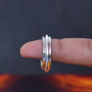 Könnte beinhalten: Ein Ring aus poliertem Silber mit einem dreifachen Banddesign. Der Ring besteht aus drei glatten, miteinander verbundenen Metallbändern, die ein geschichtetes Aussehen erzeugen. Die Silberoberfläche ist reflektierend und zeigt Lichtreflexe.