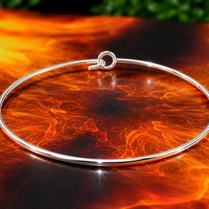 Może przedstawiać: Cienka, okrągła srebrna bransoletka typu bangle o polerowanym, odblaskowym metalicznym wykończeniu. Bransoletka ma prosty design z małym, zintegrowanym okrągłym mechanizmem zapięcia typu pętla umieszczonym na górze pośrodku.