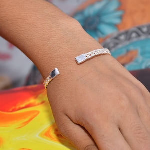 Könnte beinhalten: Ein silberfarbenes, offenes Armband mit einer geflochtenen Metallstruktur entlang des Bandes und flachen, rechteckigen Endkappen. Das Armband wird am Handgelenk getragen, und ein Ring mit einem großen, runden, weißen Stein ist am Finger sichtbar.