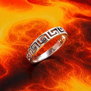 May include: A silver-toned metal ring featuring a continuous embossed Greek key pattern around the outer band. The ring has a polished finish with darkened recessed areas within the geometric design, creating a high-contrast look for the accessory.