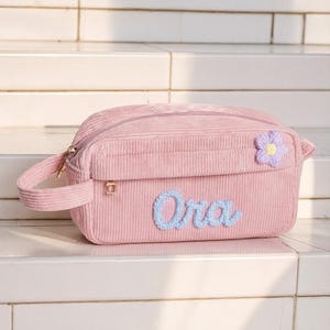 以下が含まれることがあります： サイドハンドルと上部ファスナーが付いた、ライトピンクのコーデュロイ製ポーチ。前面にはライトブルーで刺繍された「Ora」という名前と、右上に小さな紫と黄色の花のアップリケがあしらわれています。