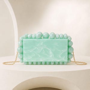 Peut inclure: Une pochette rectangulaire vert menthe avec une finition marbrée sur le panneau avant. Les bords du sac sont ornés d'une bordure de perles sphériques vert menthe de tailles variées. Une fine chaîne en métal doré est fixée de chaque côté.
