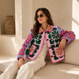 May include: A quilted jacket featuring a light pink base with vibrant floral embroidery in shades of green, red, and blue. The jacket has a collared neckline, long sleeves, two front patch pockets, and dark brown buttons down the front closure.