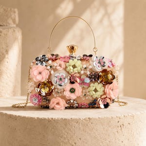 May include: A rectangular clutch bag in a soft beige tone, adorned with an array of multi-coloured fabric flowers, sequins, and faux pearls. It features a gold-toned metal top handle, a matching detachable chain strap, and a decorative gold clasp.