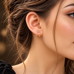Puede incluir: Un ear cuff dorado con un diseño de alambre cuadrado y extremos pequeños redondeados. La pieza se coloca en el borde exterior del cartílago de la oreja, mostrando un acabado metálico pulido y una estructura geométrica abierta y sencilla.