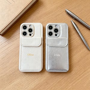 Può includere: Due custodie per smartphone con tasche per carte integrate sul retro. La custodia a sinistra è color crema con il nome Olive stampato in oro. La custodia a destra è argento metallizzato con il nome Frida stampato in bianco. Entrambe hanno fori.