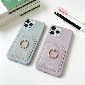 May include: Two textured phone cases in light gray and lavender, each featuring a card pocket and a gold-toned circular ring stand. The gray case has a name printed in gold, while the lavender case features the name Katie printed in white.