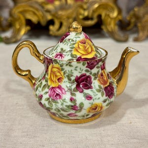 Könnte beinhalten: Eine kleine Teekanne aus Porzellan mit cremefarbenem Hintergrund und einem dichten Muster aus gelben, rosafarbenen und dunkelroten Rosen sowie grünen Blättern. Die Teekanne hat einen goldenen Griff, Ausguss, Boden und Deckelknauf sowie einen Deckel.