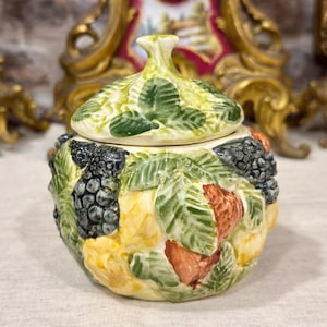 Op de afbeelding: Een keramische pot met deksel voorzien van een driedimensionaal reliëfontwerp van diverse vruchten en bessen. Het oppervlak toont gebeeldhouwde bramen, aardbeien en gele citruspartjes tussen groene bladeren, afgewerkt met een bijpassend deksel.