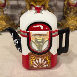 Può includere: Una teiera in ceramica a forma di jukebox vintage, caratterizzata da una combinazione di colori bianco, rosso e nero. Il beccuccio è nero con accenti di note musicali bianche, e il corpo presenta una griglia dell'altoparlante gialla, finiture argentate e un coperchio rosso.
