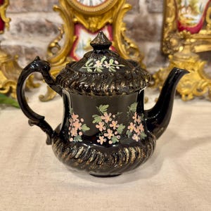 Könnte beinhalten: Eine schwarze Keramik-Teekanne mit glänzender Oberfläche, handbemalt mit rosa Blumenclustern und grünen Blättern. Die Kanne hat einen Deckel mit spitzem Knauf, einen geschwungenen Griff und einen Ausguss, verziert mit goldenen Akzenten.