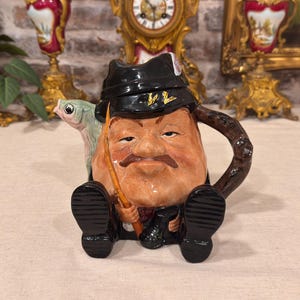 Può includere: Una teiera in ceramica a forma di persona con cappello e stivali neri, che tiene una canna da pesca con un pesce verde sul lato. La figura ha un viso abbronzato con baffi marroni e il manico è progettato per assomigliare a un ramo.