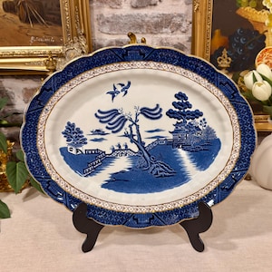 May include: An oval ceramic platter featuring a blue and white willow pattern. The central design depicts a landscape with a bridge, trees, and two birds in flight. The rim has a dark blue border with a geometric pattern and gold accents.