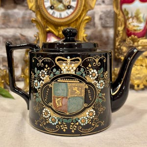 Może przedstawiać: Czarny ceramiczny imbryk z pamiątkowym wzorem upamiętniającym rekordowe panowanie królowej Wiktorii w latach 1837-1897. Z przodu znajduje się centralny herb z koroną, motywy kwiatowe w kolorze złotym, zielonym i białym oraz napis "RECORD REIGN" i "QUEEN VICTORIA".