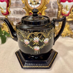 Può includere: Una teiera in ceramica nera con base esagonale abbinata, caratterizzata da un motivo in filigrana dorata e decorazioni floreali dipinte a mano nei toni del rosa e del bianco. Presenta un beccuccio curvo, un manico robusto e un coperchio con pomello.