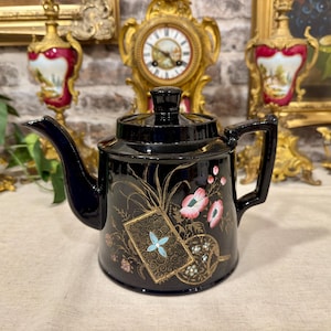 Pode incluir: Um bule de cerâmica preta brilhante com um design pintado à mão de flores cor-de-rosa, folhagem verde e um motivo retangular com contorno dourado e uma flor azul de quatro pétalas. O bule tem uma tampa preta a condizer e uma pega curva.