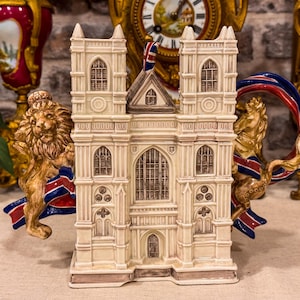 Könnte beinhalten: Eine cremefarbene Keramik-Teekanne in Form der Westminster Abbey, mit einem skulptierten Löwen auf der linken und einem Pferd auf der rechten Seite, beide drapiert in rot-weiß-blauen Union-Jack-Flaggen. Das Bauwerk zeigt detaillierte gotische Fenster.