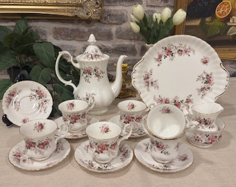 Könnte beinhalten: Ein Teeservice aus weißem Porzellan mit einem zarten rosa Rosenmuster und Goldrand. Das Set besteht aus einer Teekanne mit Deckel, einer großen Servierplatte, einem Milchkännchen sowie mehreren Teetassen mit passenden Untertassen.