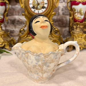 May include: A ceramic creamer jug featuring a sculpted figure of a person with black hair and orange lips emerging from the top. The jug has a ruffled rim and a handle, decorated with a mottled beige and light blue speckled pattern.