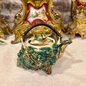 Op de afbeelding: Een keramische theepot in de vorm van een schelp met een glanzend groen en bruin glazuur. De theepot heeft een textuurrijk oppervlak, een bijpassend deksel met een kleine schelpknop, een gebogen tuit en een zwart omwikkeld draadhandvat.
