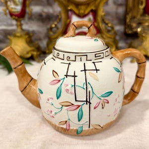Op de afbeelding: Een crèmekleurige keramische theepot met een textuur op het oppervlak, voorzien van een bruin handvat, tuit en dekselknop in bamboestijl. De behuizing is versierd met geschilderde roze en blauwgroene bladeren en zwarte geometrische lijnen.