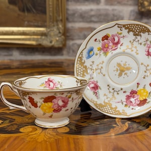 Op de afbeelding: Een witte porseleinen theekop en bijpassende schotel met een kleurrijk bloemmotief van roze, gele en rode bloemen, geaccentueerd door gedetailleerde gouden filigraan langs de geschulpte randen en een goudkleurig handvat en voet.