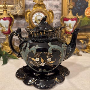 Könnte beinhalten: Eine schwarze Keramik-Teekanne mit passender Untertasse, verziert mit goldfarbenen Blumenmotiven, salbeigrünen Schnörkeln und gewellten Rändern. Die Teekanne hat einen geschwungenen Griff, einen spitzen Ausguss und einen Deckel mit Zierknauf.