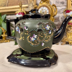 Peut inclure: Une théière vert olive mat avec un couvercle, une anse, un bec et une base noir brillant. Le corps présente des motifs dorés tourbillonnants peints à la main et de petites fleurs bleues et violettes. La théière repose sur une soucoupe assortie.