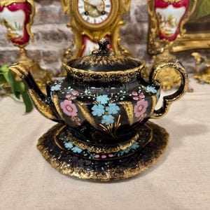 Könnte beinhalten: Eine schwarze Keramik-Teekanne mit passender Untertasse in einem gewundenen, gerippten Design. Das Set ist mit handgemalten hellblauen und rosafarbenen Blumenmotiven verziert und mit einer dicken Goldverzierung an den Rändern, dem Griff und dem Ausguss versehen.