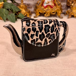 Op de afbeelding: Een vierkante theepot met een glanzend zwarte basis en een beige klep met een zwart luipaardpatroon. De theepot heeft een metallic zilveren tuit en handvat, met de initialen AR in het wit gedrukt in de rechterbenedenhoek van het zwarte deel.